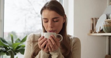 Güzel bir kadın evdeki bardaktan sıcak bir içecek içerken pencereden dışarı bakıyor.