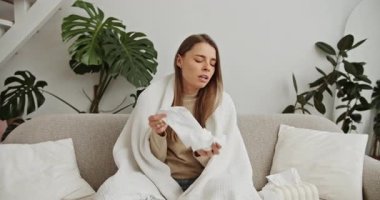 Sağlıksız bir kadın battaniyeye sarılıp evdeki kanepede oturuyor, grip ya da soğuk algınlığından muzdarip.