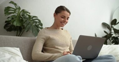 Genç ve güzel bir kadın rahat bir oturma odasında laptopuyla çalışırken kanepede dinleniyor.