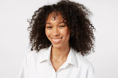 Genç kadın doğal kıvırcık saçlarıyla gülümsüyor, beyaz arka planda mutluluk ve neşe ifade ediyor.