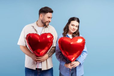 Mavi arka planda izole edilmiş kameraya bakan kırmızı balonları tutan mutlu çiftin portresi. Sevgililer Günü konsepti