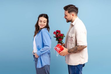 Yakışıklı romantik adam mavi arka plandaki güzel kıza kırmızı çiçekler ve kırmızı kutu hediye ediyor. Tatil, aşk, ilişki, Sevgililer Günü konsepti