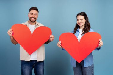 Kırmızı kalp sevgililer günü kartı taşıyan gülümseyen çift mavi arka planda izole edilmiş kameraya bakıyor. Sevgililer Günü konsepti
