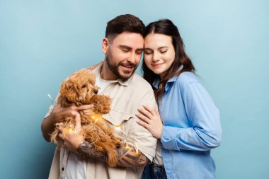Aşık mutlu bir çift, yakışıklı bir adam ve neşeli bir kadın küçük sevimli bir köpek tutuyor, mavi arka planda soyutlanmış bir şekilde sarılıyorlar. Hayvan konseptine bayılırım.