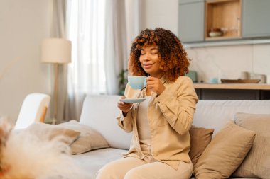 Genç bir kadın kanepede oturuyor, sıcak bir içecek kokluyor, evinde rahatlık ve rahatlık buluyor.