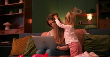 Küçük kızı saçlarıyla oynarken evde dizüstü bilgisayarla çalışan meşgul bir anne.