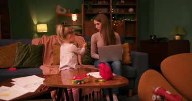 Oyuncu kızı serbest çalışan annesini rahatsız ediyor. Oturma odasında dizüstü bilgisayarla çalışıyor.