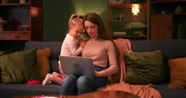 Çalışan bir kadın dizüstü bilgisayar kullanırken küçük kızı kanepesinde oturmuş onu izliyor.