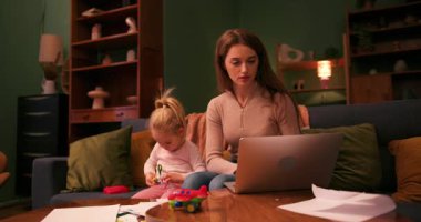 Küçük kızı kanepede oynarken evde dizüstü bilgisayarda çalışan genç bir iş kadını.