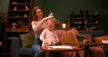 Küçük kız kâğıdı makasla keserken, annesi bebeğinin saçlarını şekillendiriyor.