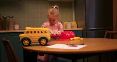 Pijamalı küçük bir kız mutfak masasında sarı oyuncak bir otobüs ve kırmızı plastik bir miğferle oynuyor.