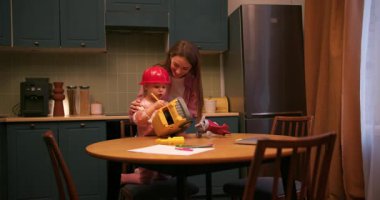 Genç bir kadın ve kasklı bir bebek mutfak masasında oyuncak bir çöp kamyonuyla oynuyorlar.