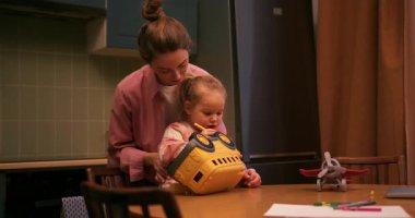 Anne, kızının saçlarını şekillendirirken çocuk mutfak masasında oyuncak bir otobüsle oynuyor.