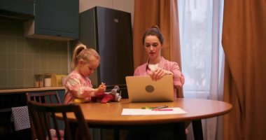 Küçük kızı mutfak masasında resim çizerken genç bir anne dizüstü bilgisayarla uzaktan çalışıyor.