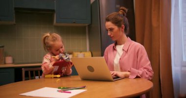 Küçük kızı aynı masada oyuncaklarla oynarken genç bir kadın evde dizüstü bilgisayarla çalışıyor.