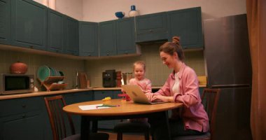 Çalışan bir anne, mutfak masasında kızıyla birlikte çalışıyor. Uzaktan kumandalı iş ve aileyi dengeliyor.