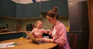 Çalışan anne dizüstü bilgisayarda çalışırken neşeli kızı mutfak masasında pastel boyalarla resim çiziyor.