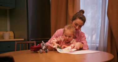 Genç anne, küçük kızına mutfak masasında otururken resim çizmesinde yardım ediyor.