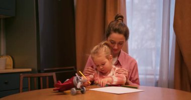 Genç bir kadın, kızına, akşamları tahta bir masada renkli kalemlerle resim çizmesinde yardım ediyor.