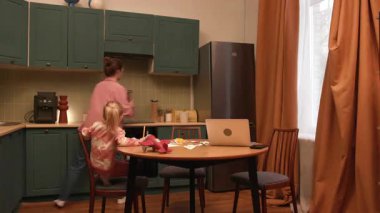 Çalışan anne dizüstü bilgisayarına odaklanmaya çalışırken kızı mutfakta ilgi istiyor.