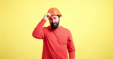 Sakallı erkek müteahhit sert bir şapka takıyor ve parlak sarı bir arka planda güvenle gülümsüyor.