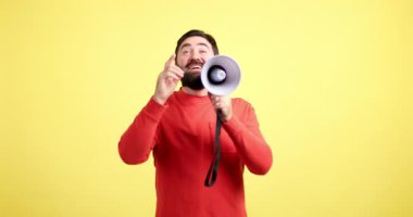 Kırmızı kazaklı olumlu sakallı adam sarı arka planda megafonla bir duyuru yapıyor.
