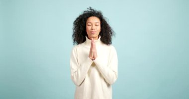 Genç Afrikalı Amerikalı kadın ellerini mavi bir arka plana bağlayıp dua ediyor.