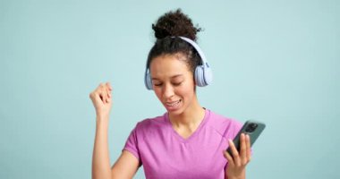 Kulaklıklı, müzik dinleyen ve mavi arka planda akıllı telefonuyla dans eden genç bir kadın.