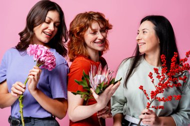 Farklı çiçeklerle gülümseyen üç kadın, çeşitliliği, dostluğu ve mutluluğu simgeliyor.