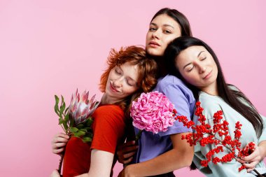 Üç farklı kadın birbirine yakın duruyor, beraberlik gösteriyorlar ve farklı çiçek türleri tutuyorlar.