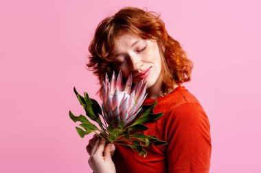 Çilli bir kadın, büyük pembe bir çiçeğe sarılıyor ve gözleri kapalı.