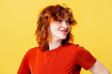 Kızıl saçlı, çilli, gülümseyen, diş teli takan, sarı arka plana bakan genç bir kadın.