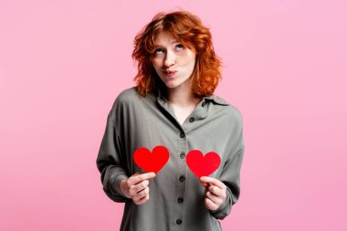 Kızıl saçlı kadın pembe arka planda iki kırmızı kağıt kalp tutuyor. Romantik düşüncelerini dile getiriyor.