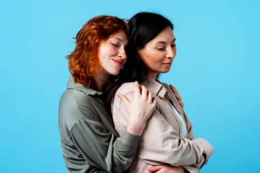 Sarılan, şefkat gösteren, dostluk gösteren ve mavi arka planda samimi bir bağ kuran genç kadınlar.