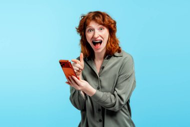 Diş teli takmış, şaşırmış ve heyecanlanmış genç bir kadın cep telefonu tutuyor ve içerikle meşgul oluyor.