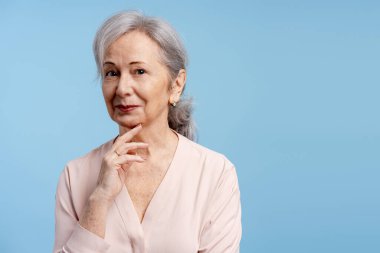 Güzel, gülümseyen, olgun, gri saçlı bir kadın mavi arka planda izole bir şekilde poz veriyor. Kameraya bakıyor, mekanı kopyalıyor. Reklam kavramı