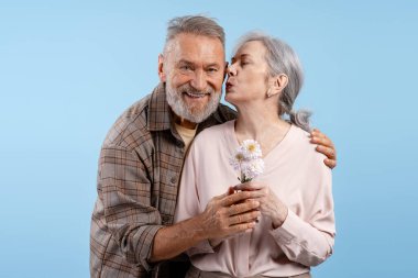 Yakışıklı, gülümseyen, yaşlı bir adam mutlu olgun bir kadına pembe çiçek veriyor, o da onu mavi arka planda öpüyor. Aşk, ilişki kavramı