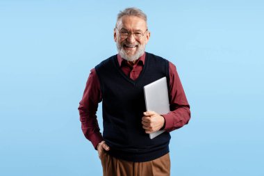 Gülümseyen, gri sakallı, gözlüklü, dizüstü bilgisayar takan, mavi arka planda izole edilmiş kameraya bakan adam. Çevrimiçi teknoloji kavramı