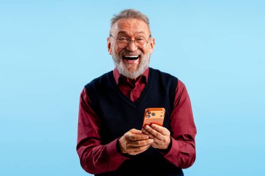 Heyecanlı, gülümseyen son sınıf öğrencisi cep telefonu tutuyor, mavi arka planda izole edilmiş kameraya bakarak mobil uygulama kullanıyor. Çevrimiçi teknoloji kavramı