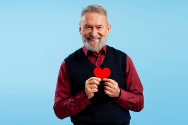 Mutlu gülümseyen son sınıf öğrencisi elinde kırmızı kalp sevgilisiyle mavi arka planda izole edilmiş kameraya bakıyor. Sevgililer Günü konsepti