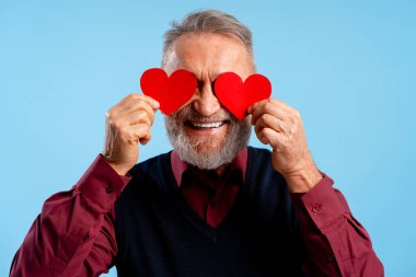 Kırmızı kalp taşıyan mutlu, pozitif yaşlı adam mavi arka planda izole edilmiş poz veriyor. Sevgililer Günü konsepti