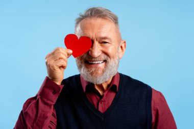 Mavi arka planda izole edilmiş kameraya bakarak kırmızı kalp tutan yakışıklı, son sınıf öğrencisi. Aşk, Sevgililer Günü konsepti