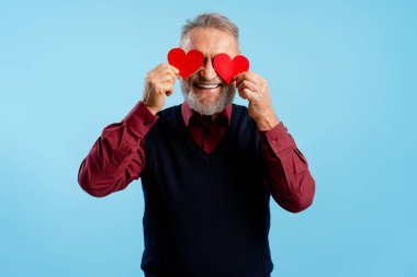 Gülümseyen mutlu, pozitif yaşlı adam elinde kırmızı kalplerle mavi arka planda yalnız poz veriyor. Sevgililer Günü konsepti
