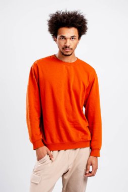 Turuncu kazaklı ve bej pantolonlu afro saçlı genç adam kameraya bakıyor.