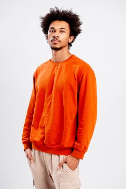 Kendine güvenen genç bir adam, günlük turuncu bir kazak ve bej bir pantolon giyiyor.