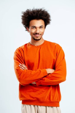 Turuncu kazaklı afro saçlı genç bir adam stüdyo arka planında güvenle gülümsüyor.