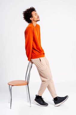 Turuncu kazaklı ve bej pantolonlu genç adam sandalyeye yaslanmış, odaklanmış bir şekilde yukarı bakıyor.