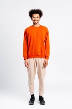 Kıvırcık saçlı genç adam gülümsüyor, beyaz arka planda kendine güvenen bir stüdyoda duruyor.