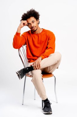 Genç adam gündelik kıyafetler giyiyor, bir sandalyeye oturuyor, kendinden emin ve rahat bir tavır sergiliyor.