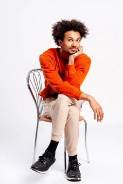 Turuncu süveter giyen genç bir adam sandalyeye oturmuş çenesi çenesinde kameraya bakıyor.